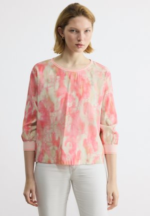 Femme portant un chemisier à manches longues rose et beige à motif, avec des manches retroussées, et un pantalon blanc, debout devant un fond uni.