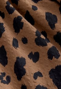Brown fabric with a textured surface featuring irregular black spots. The pattern has varying sizes and shapes, creating a bold contrast.