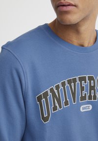 Sudadera azul con escote redondo, que presenta una impresión en blanco y negro de "UNIVERSITY" y el año "MMXX" en un emblema redondeado. Textura suave.