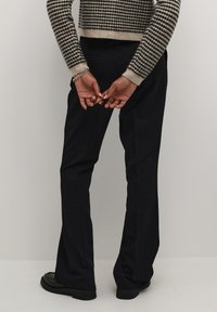 Sorte flared bukser med en glat tekstur, parret med en cropped, mønstret sweater i beige og sort. Hænderne foldet bag ryggen.