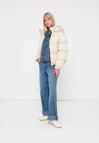 Puffjacka i krämfärg med huva, tillsammans med en blå denimjacka och vida jeans. Vita sneakers fulländar outfiten.