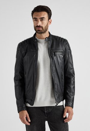 Zwarte leren jas met geribbelde schouderdetails, ritssluitingen en een staande kraag. Gedragen over een grijze trui en donkere jeans.