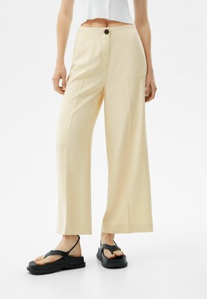 Vrouw draagt beige wijde broek met hoge taille en zwarte plateauzandalen, staand tegen een witte achtergrond.