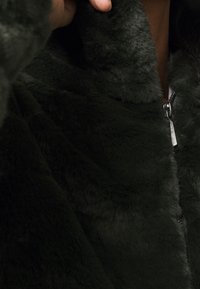 Ciemnozielona pluszowa kurtka zapinana na zamek, o puszystej fakturze, z wysokim kołnierzem i srebrnym detalem zamka z przodu.