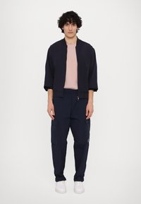 Mornarska bombarder jakna in cargo hlače, kombinirane z svetlo roza majico. Teksturirana tkanina, detajli z zadrgo in žepi na hlačah. Bele športne superge.