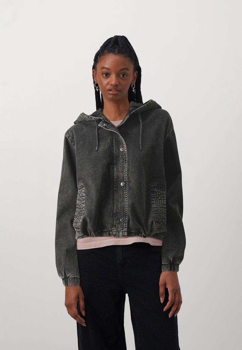 Chaqueta de mezclilla negra con capucha, cierre de botones en la parte delantera, bolsillos laterales, puños elásticos y cintura ajustada. Presenta una textura desgastada.