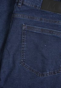 Jeans de mezclilla azul oscuro con un diseño clásico de cinco bolsillos, costuras visibles y una etiqueta de cuero con una letra M en relieve.