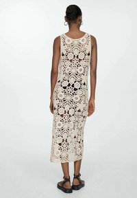 Abito in crochet beige con motivi floreali, senza maniche, di lunghezza media, con lavorazione a rete e vestibilità rilassata, abbinato a sandali neri.