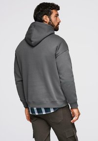 Mężczyzna z ciemnymi włosami i brodą, noszący szary hoodie na zielono-białą koszulę w kratę oraz ciemne spodnie cargo, zwrócony plecami na jednolitym tle.