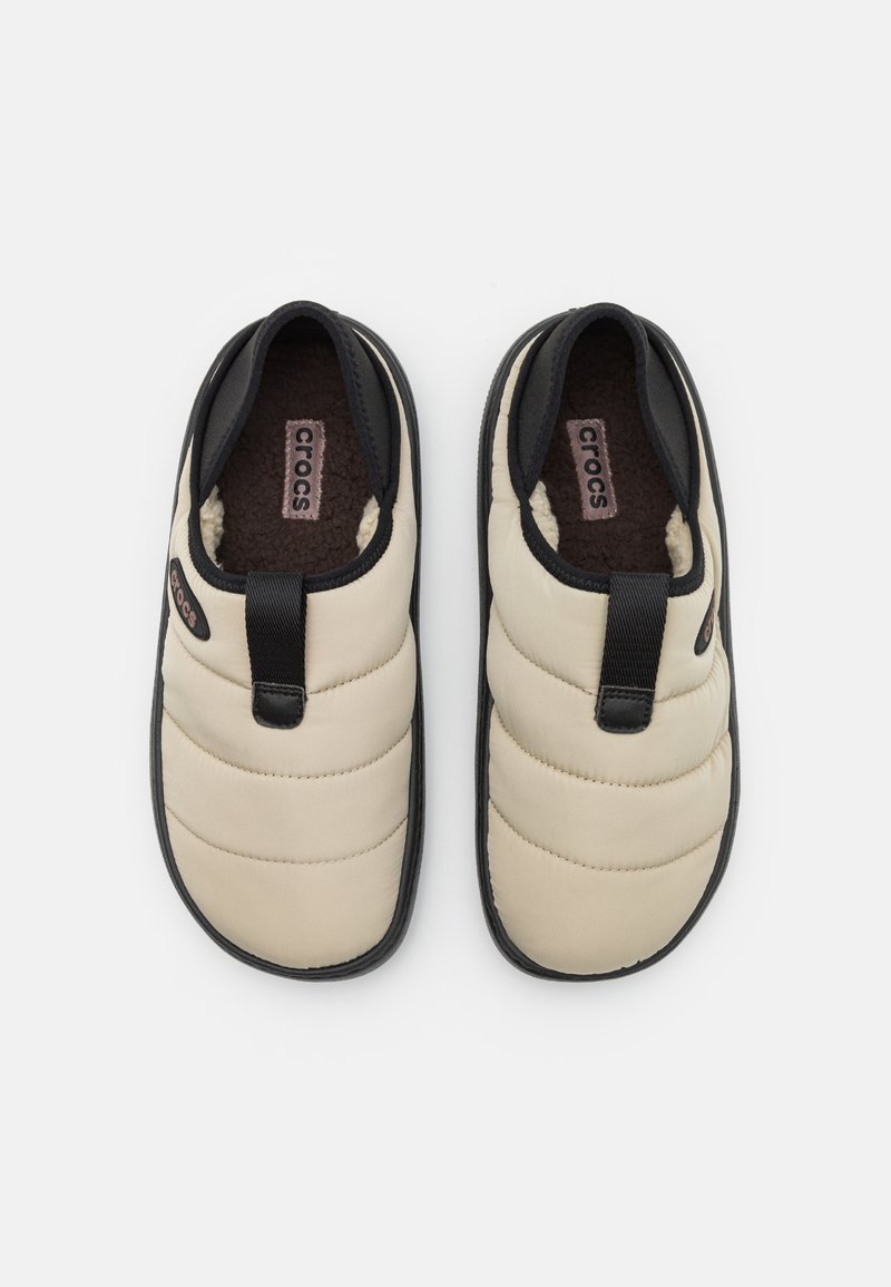 Zapatillas deslizables beige con diseño acolchado, detalles negros, forro suave y una lengüeta. Suela de goma texturizada para mayor agarre.