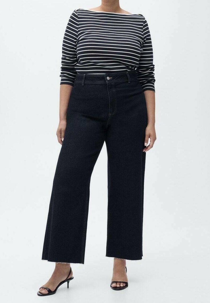 Femme portant un haut à manches longues rayé noir et blanc, un jean noir taille haute à jambes larges et des talons ouverts noirs, debout sur un fond blanc.