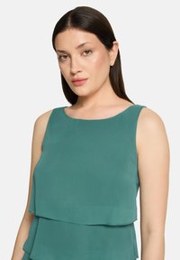 Haut sans manches de couleur sarcelle, présentant un design en couches avec une texture lisse. Encolure arrondie et coupe décontractée. Aucun motif ni détail métallique visible.