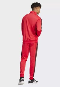 Tuta rossa realizzata in tessuto liscio con strisce laterali nere. Include una giacca con zip e pantaloni affusolati. Indossata con sneakers nere e bianche.