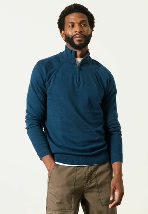 Mann mit Bart, der einen blauen Quarter-Zip-Pullover und braune Hose trägt, steht mit einer Hand in der Tasche vor einem schlichten hellen Hintergrund.