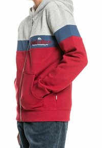 Persona che indossa una felpa con zip, con blocchi di colore grigio, blu e rosso e il logo "Quiksilver Original Boardriding Co." sul petto.