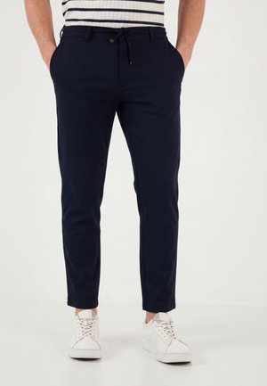 Personne portant un pantalon slim bleu marine avec cordon et des baskets blanches sur un fond clair uni.