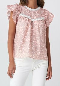 Ärmellose pinke Bluse mit weißen abstrakten Punkten, Rüschencapärmel, Spitzenbesatz am Ausschnitt, getragen mit weißen Hosen.