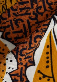 Tissu présentant un design vibrant et complexe avec des tons orange et jaune, des détails noirs et un tissage texturé. Les motifs incluent des formes abstraites et des points.