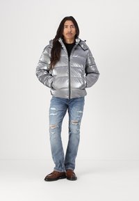 Giacca a vento argento con cuciture orizzontali, chiusura con zip e cappuccio; abbinata a jeans blu strappati e scarpe marroni.
