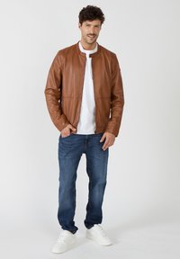 Braune Lederjacke mit Stehkragen, Reißverschluss und zwei Seitentaschen, kombiniert mit blauen Jeans und weißen Sneakers.