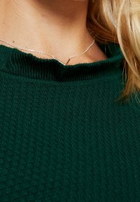 Zbliżenie osoby noszącej delikatny srebrny naszyjnik z małym wisiorkiem, teksturowany ciemnozielony sweter oraz długie blond włosy.