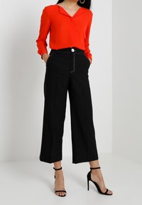 Blouse rouge à col en V et manches longues, associée à un pantalon large noir avec bouton et surpiqûres contrastées, portée avec des talons noirs.