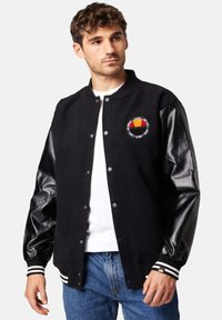 Black varsity jacket with leather sleeves, embroidered patch on the chest, ribbed knit cuffs with white stripes, and snap button closure.
