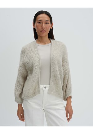 Frau mit glatten dunklen Haaren trägt eine beige strukturierte Strickjacke über einem gerippten Oberteil und hoch taillierten weißen Hosen und steht vor einem schlichten Hintergrund.