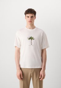 Balta medvilninė marškinėliai su žalia palmė grafika ant priekio, trumpomis rankovėmis ir apvalia iškirpte. Modelis vilki smėlio spalvos kelnes.
