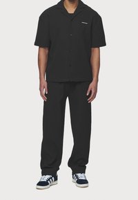 Black short-sleeve shirt with a collar and button closure, paired with matching loose-fitting black pants and navy sneakers with white stripes.