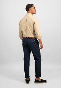 Beige overhemd met lange mouwen en opgerolde mouwen, marineblauwe broek, geweven riem en zwarte espadrilles, gezien vanaf de achterkant.