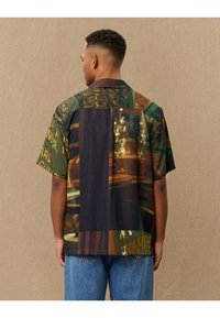Short-sleeve shirt featuring a multicolored print with dark tones, text elements, and varying textures in a loose, relaxed fit.