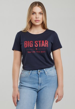 Jauna blondinė moteris dėvi tamsiai mėlyną marškinėlį su raudonu užrašu „BIG STAR Your Life. Your Jeans.“ ir šviesiai mėlynas aukšto liemens džinsus.