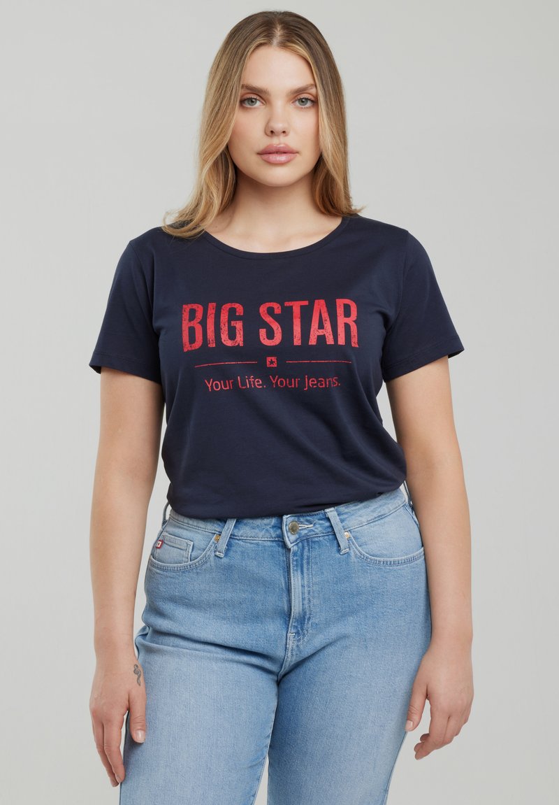 Jauna blondinė moteris dėvi tamsiai mėlyną marškinėlį su raudonu užrašu „BIG STAR Your Life. Your Jeans.“ ir šviesiai mėlynas aukšto liemens džinsus.