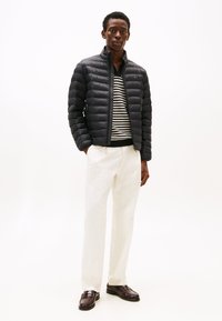 Veste matelassée noire à motif matelassé horizontal, associée à un pull à rayures, un pantalon crème et des chaussures en cuir marron.