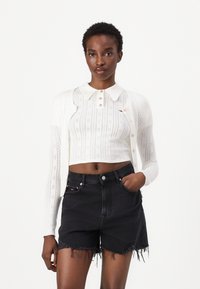Woman wearing a white knit cropped cardigan and polo shirt with black high-waisted frayed denim shorts, standing against a plain background.