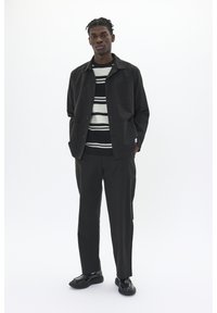Black jacket with front pockets, worn over a striped sweater in black, white, and gray; paired with black trousers and shoes.
