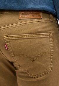 Πίσω τσέπη και δερμάτινο ταμπελάκι μάρκας σε καφέ τζιν Levi's 511 που φοριούνται με μπλε πουκάμισο περασμένο μέσα. Μέγεθος W31 L34.