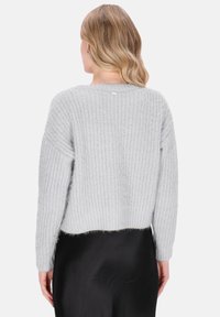 Kobieta z falującymi blond włosami, ubrana w jasnoszary sweter dzianinowy i czarną spódnicę, widoczna od tyłu na jednolitym tle.