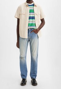 Camisa corta de manga beis claro sobre una camiseta verde a rayas, combinada con jeans azul deslavado que presentan un desgarro y zapatos negros.