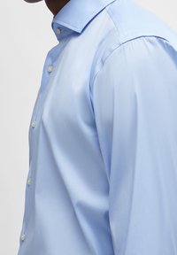 Camisa azul claro de tela suave, con cuello puntiagudo y botones en la parte delantera, con costuras visibles en los hombros y corte entallado.