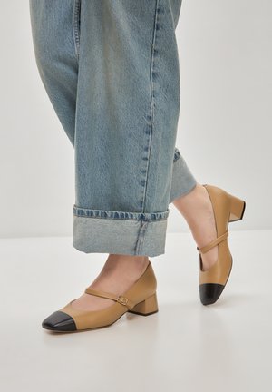 Scarpe Mary Jane in pelle marrone con punta nera, tacco basso e cinturino alla caviglia, abbinate a pantaloni di jeans leggeri blu chiaro risvoltati.