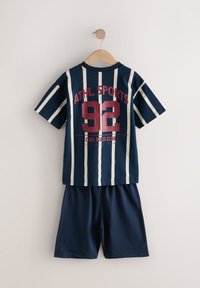 Chemise en coton bleu marine à manches courtes avec rayures blanches ; comporte un texte rouge "ATHL. SPORTS 92" et un short bleu marine assorti, suspendus à un crochet en bois.