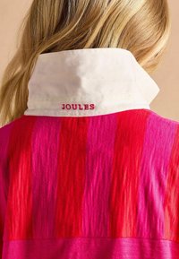 Košeľa s golierom v ružovom a červenom vertikálnom pruhu, so bielym golierom, na ktorom je na zadnej strane vyšívané slovo "JOULES" červenou niťou.