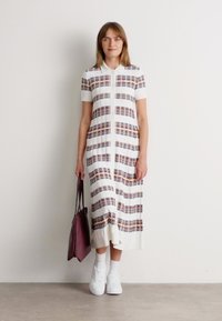 Γυναίκα που φοράει ένα λευκό midi φόρεμα με οριζόντιες καρό ρίγες, λευκά αθλητικά παπούτσια, λευκές κάλτσες και κρατάει μια μπορντό τσάντα.