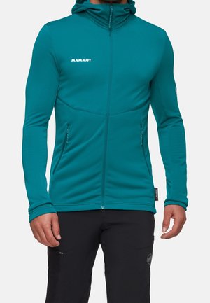 Man wearing teal full-zip hooded jacket with zipped pockets and black pants, shown from neck to hips.