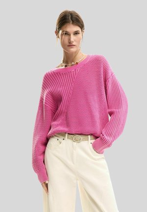 Žena oblečená v textúrovanom ružovom pletenom svetri s dlhými rukávmi a krémových širokých nohaviciach stojaca pred jednofarebným sivým pozadím.