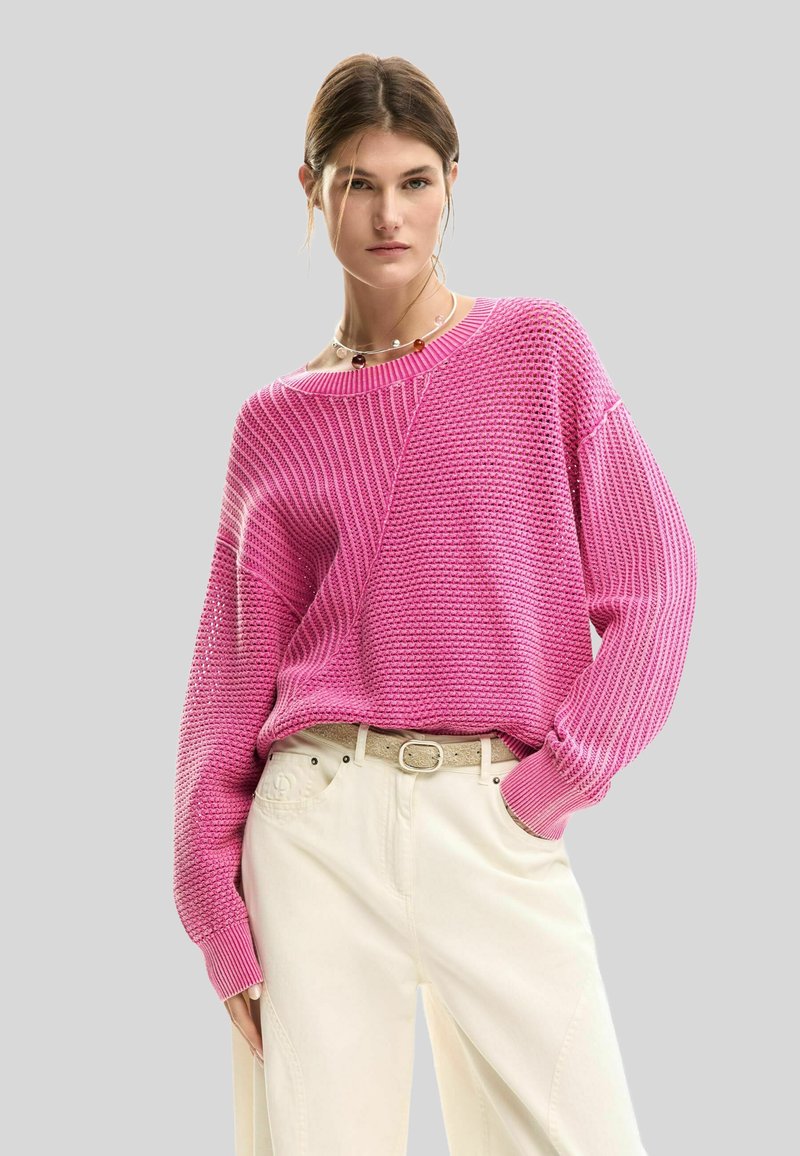 Donna che indossa un maglione rosa lavorato a maglia con maniche lunghe e pantaloni crema a gamba larga, in piedi contro uno sfondo grigio uniforme.