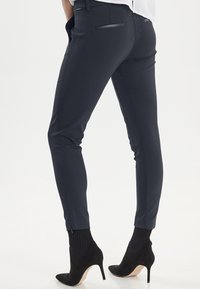 Tumši pelēki pielāgotie bikses ar elegantiem sānu paneļiem, kontrastējošām aizmugurējām kabatām un rāvējslēdzēja detaļu hemā. Apvienoti ar melnām zābakiem ar papēdi.