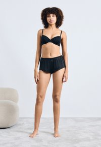 Zwarte gevoerde bh en bijpassende zwarte shorts, gemaakt van een gladde stof. Losvallende shorts met een elastische tailleband, model staat op blote voeten.